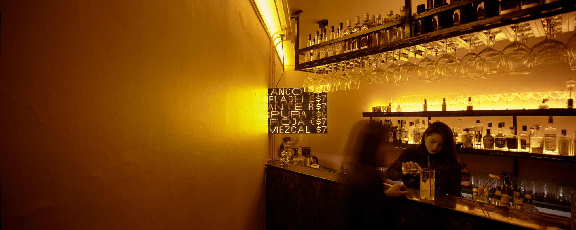 A small screen with drink prices behind a bar while a bartender pours a drink.